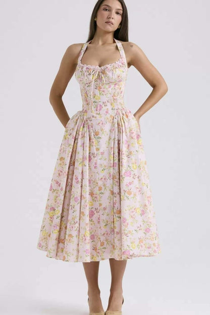 Pink - A woman with her hair in an updo smiles as she twirls in a sunlit garden, wearing a pink floral print halterneck midi dress with a corset-style ruched bodice and a full, flared skirt.