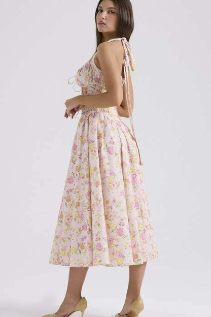 Pink - A woman with her hair in an updo smiles as she twirls in a sunlit garden, wearing a pink floral print halterneck midi dress with a corset-style ruched bodice and a full, flared skirt.