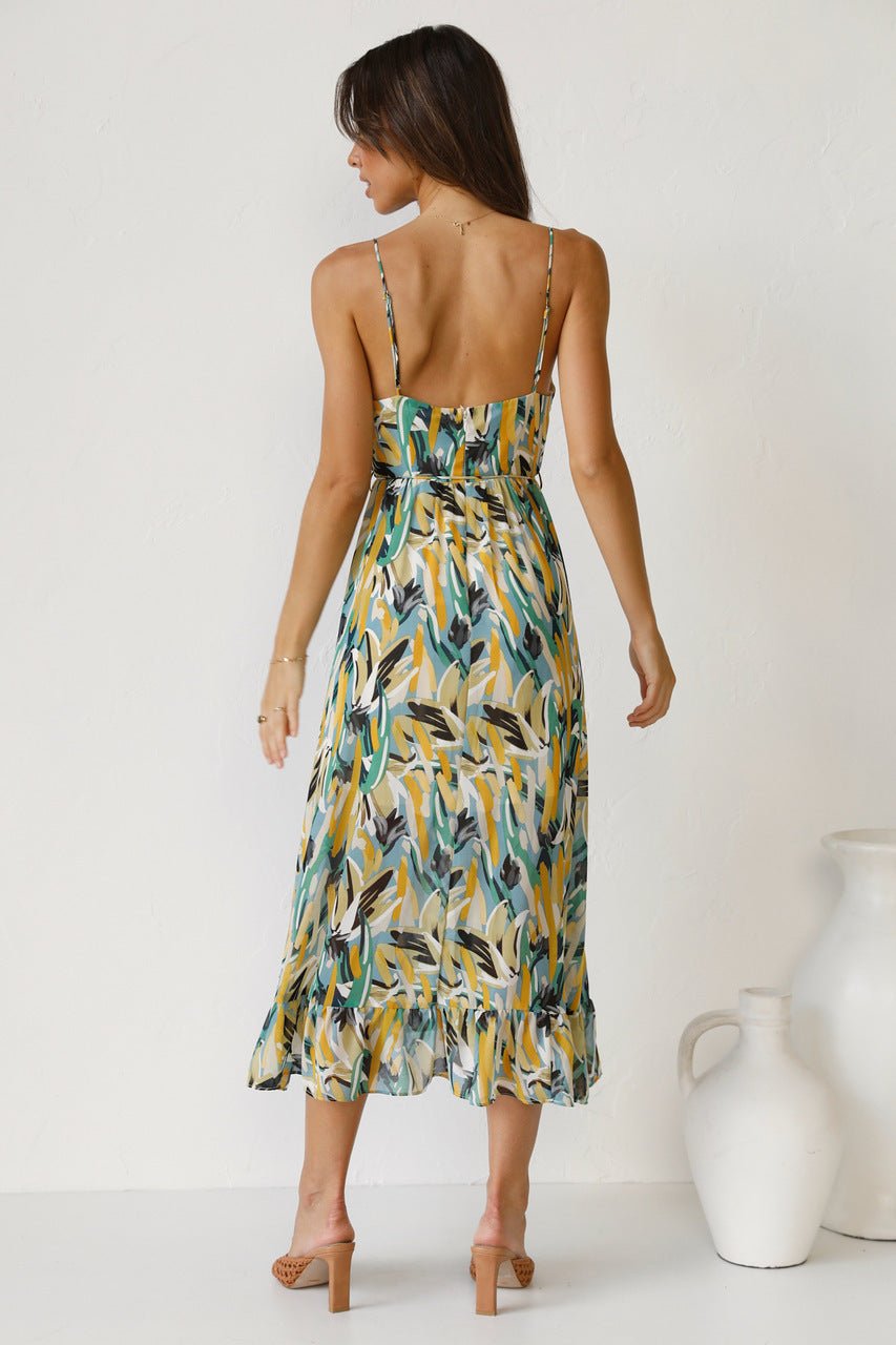 A woman with long brown hair smiles while wearing a V-neck midi dress with spaghetti straps. The dress features a yellow, green, and black abstract brushstroke print and has a ruffled hem. She is standing in a white room, wearing brown woven sandals.