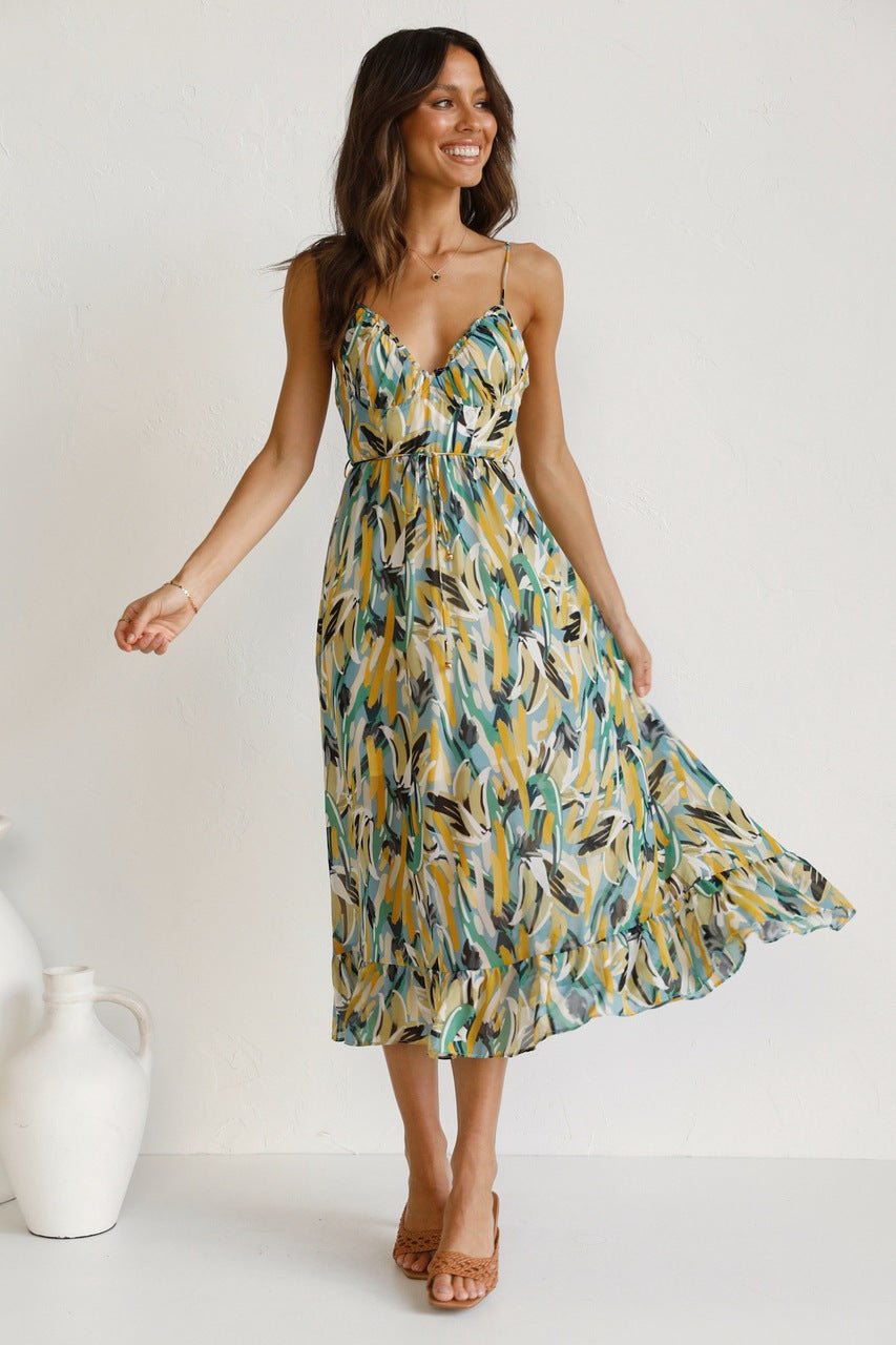 A woman with long brown hair smiles while wearing a V-neck midi dress with spaghetti straps. The dress features a yellow, green, and black abstract brushstroke print and has a ruffled hem. She is standing in a white room, wearing brown woven sandals.