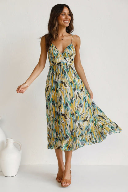 A woman with long brown hair smiles while wearing a V-neck midi dress with spaghetti straps. The dress features a yellow, green, and black abstract brushstroke print and has a ruffled hem. She is standing in a white room, wearing brown woven sandals.