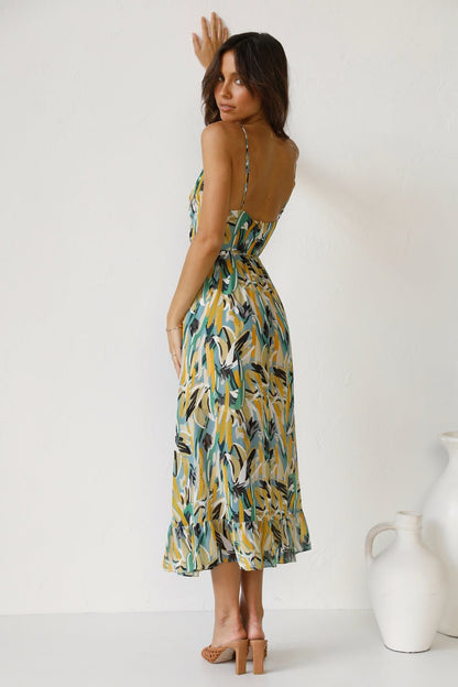 A woman with long brown hair smiles while wearing a V-neck midi dress with spaghetti straps. The dress features a yellow, green, and black abstract brushstroke print and has a ruffled hem. She is standing in a white room, wearing brown woven sandals.