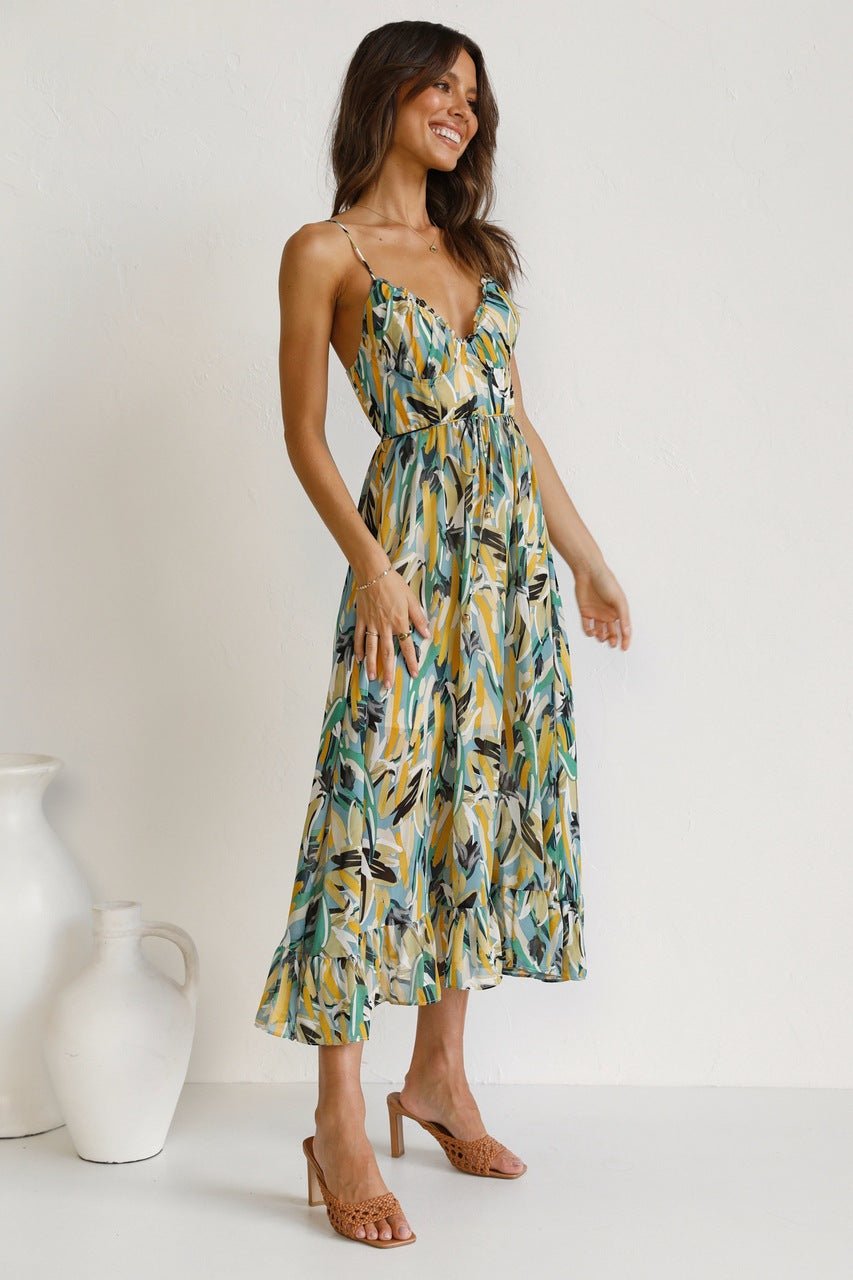 A woman with long brown hair smiles while wearing a V-neck midi dress with spaghetti straps. The dress features a yellow, green, and black abstract brushstroke print and has a ruffled hem. She is standing in a white room, wearing brown woven sandals.