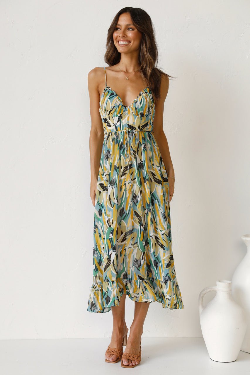 A woman with long brown hair smiles while wearing a V-neck midi dress with spaghetti straps. The dress features a yellow, green, and black abstract brushstroke print and has a ruffled hem. She is standing in a white room, wearing brown woven sandals.