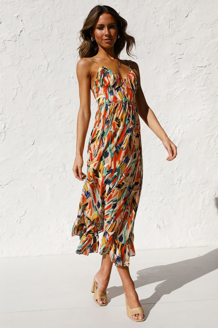 A woman with long brown hair smiles while wearing a V-neck midi dress with spaghetti straps. The dress features a yellow, green, and black abstract brushstroke print and has a ruffled hem. She is standing in a white room, wearing brown woven sandals.