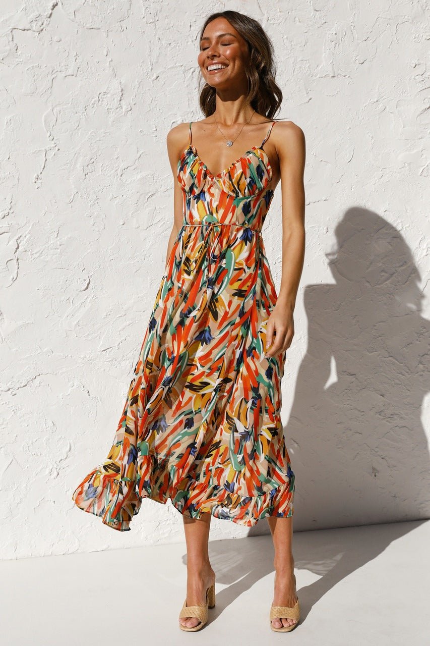 A woman with long brown hair smiles while wearing a V-neck midi dress with spaghetti straps. The dress features a yellow, green, and black abstract brushstroke print and has a ruffled hem. She is standing in a white room, wearing brown woven sandals.