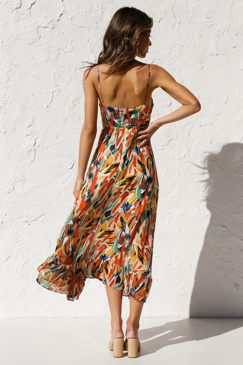 A woman with long brown hair smiles while wearing a V-neck midi dress with spaghetti straps. The dress features a yellow, green, and black abstract brushstroke print and has a ruffled hem. She is standing in a white room, wearing brown woven sandals.