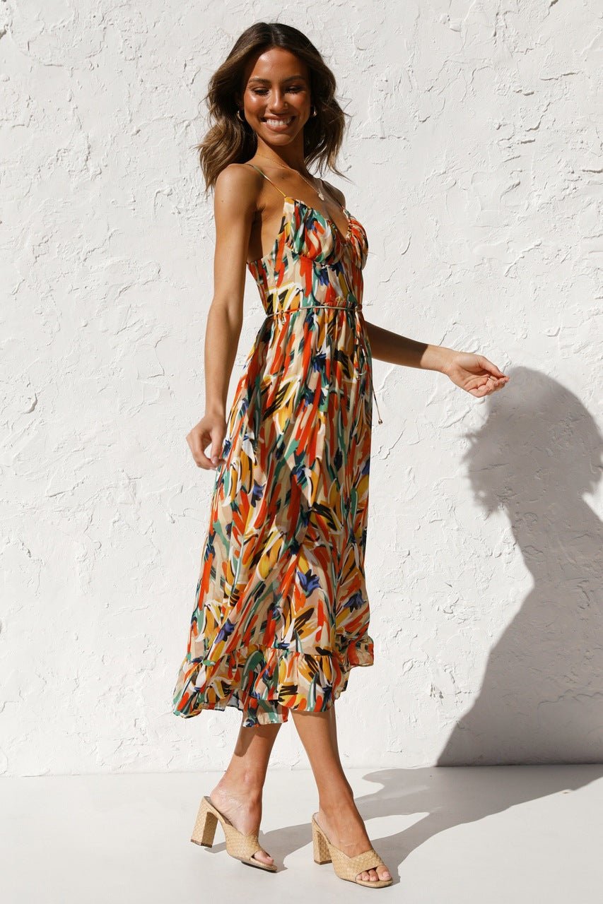 A woman with long brown hair smiles while wearing a V-neck midi dress with spaghetti straps. The dress features a yellow, green, and black abstract brushstroke print and has a ruffled hem. She is standing in a white room, wearing brown woven sandals.