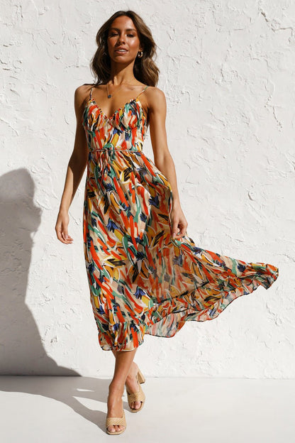 A woman with long brown hair smiles while wearing a V-neck midi dress with spaghetti straps. The dress features a yellow, green, and black abstract brushstroke print and has a ruffled hem. She is standing in a white room, wearing brown woven sandals.