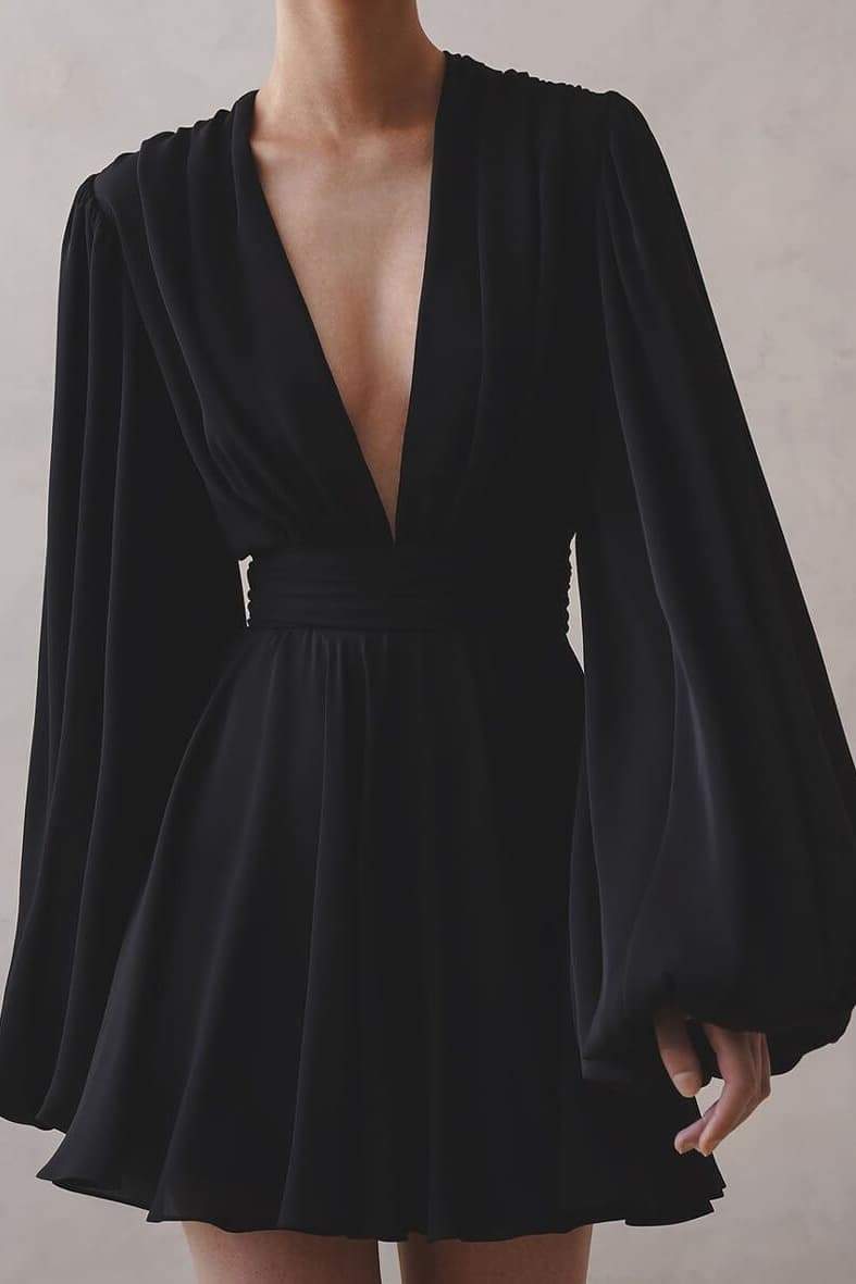 A woman with her hair in a sleek bun models a black mini dress with a plunging V-neckline, extremely voluminous long sleeves, a wide fitted waistband, and a short, flared skirt.