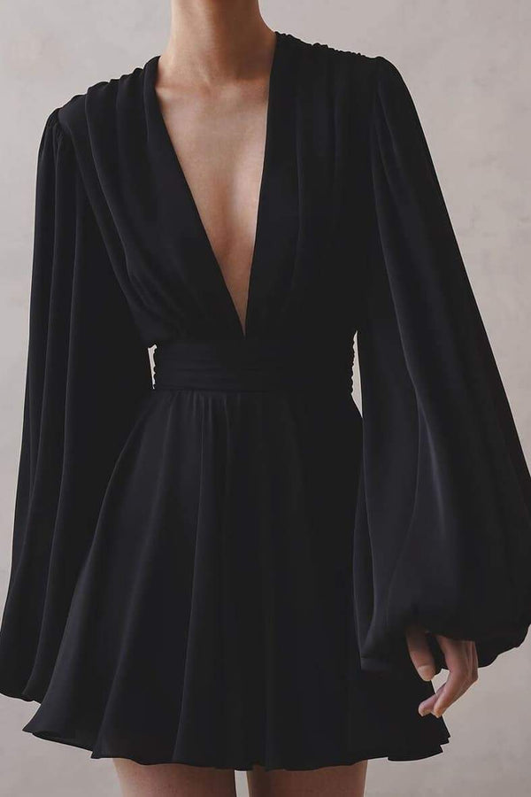 A woman with her hair in a sleek bun models a black mini dress with a plunging V-neckline, extremely voluminous long sleeves, a wide fitted waistband, and a short, flared skirt.