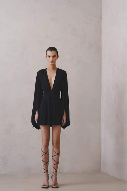 A woman with her hair in a sleek bun models a black mini dress with a plunging V-neckline, extremely voluminous long sleeves, a wide fitted waistband, and a short, flared skirt.