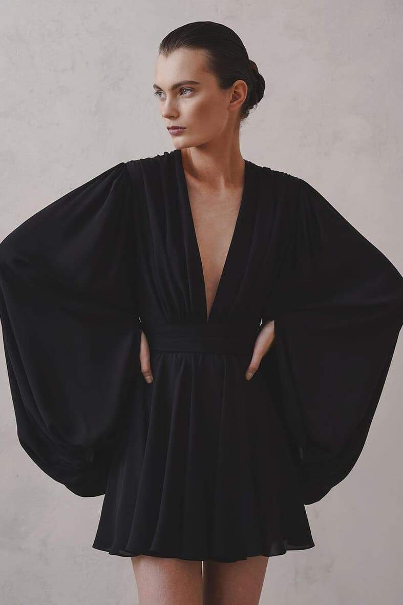 A woman with her hair in a sleek bun models a black mini dress with a plunging V-neckline, extremely voluminous long sleeves, a wide fitted waistband, and a short, flared skirt.