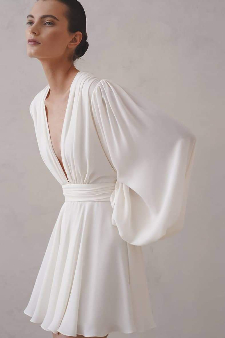 A woman with her hair in a sleek bun models a black mini dress with a plunging V-neckline, extremely voluminous long sleeves, a wide fitted waistband, and a short, flared skirt.