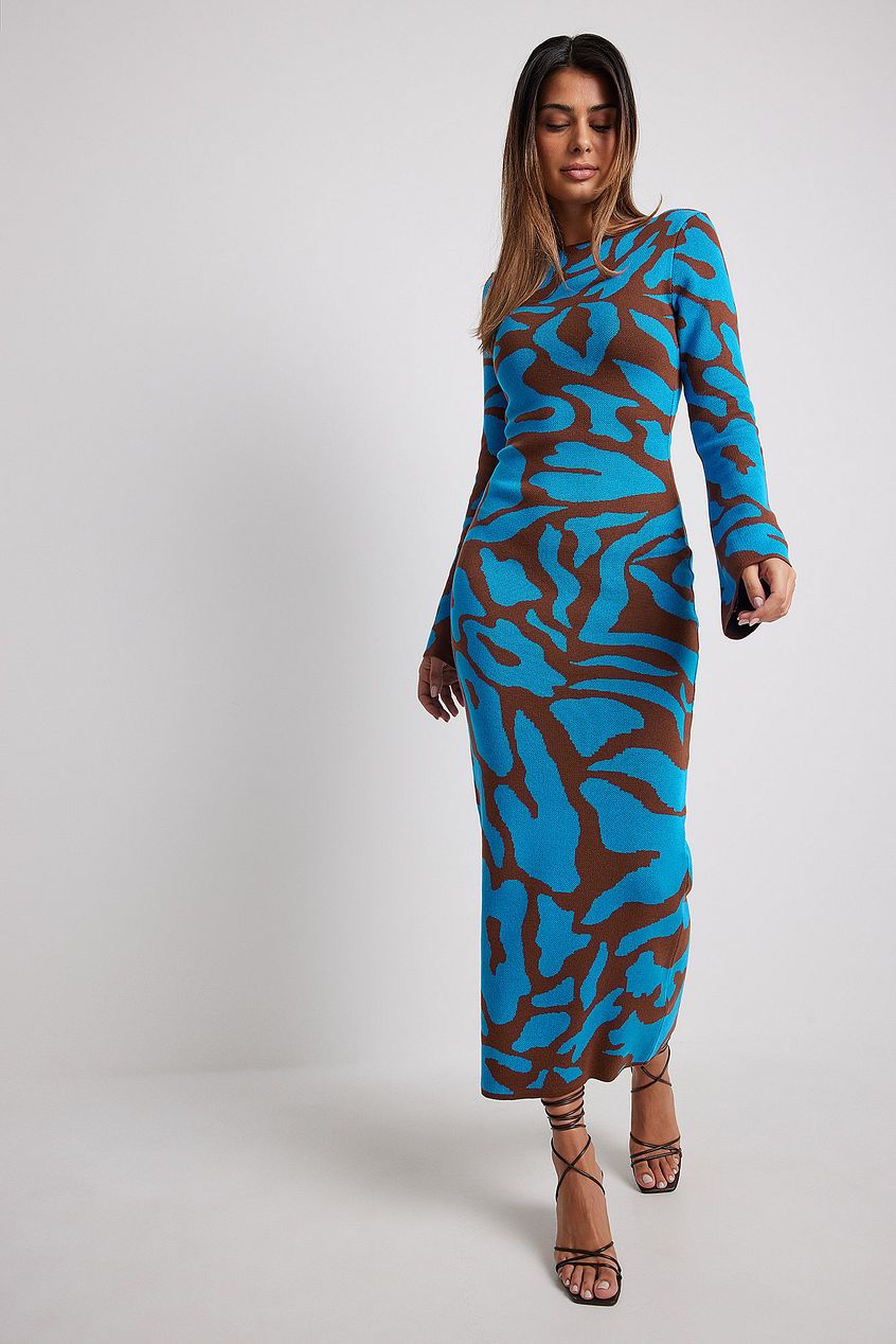 A woman models a long-sleeve, crewneck bodycon maxi dress featuring a bold abstract animal print in bright blue and brown. She is walking forward against a plain light grey background, wearing black strappy high heels.