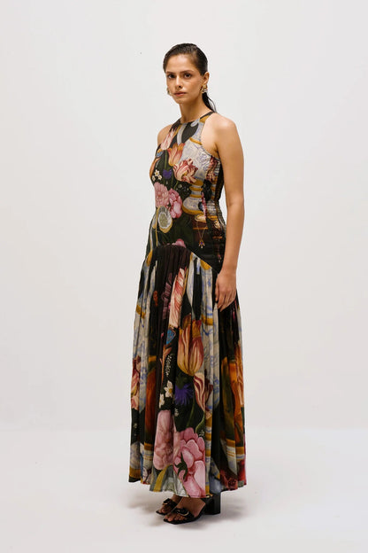 A woman with dark hair slicked back stands against a white background, wearing a full-length, sleeveless halter dress. The dress features a dark, intricate print of flowers, lemons, and antique vases, with a fitted bodice and a long, pleated A-line skirt.