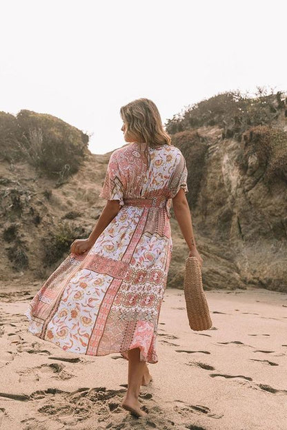 Multi - A woman on a sandy beach wearing a long, flowing pink and white bohemian print maxi dress with a deep V-neck, wide short sleeves, and a high leg slit, holding a woven straw bag.