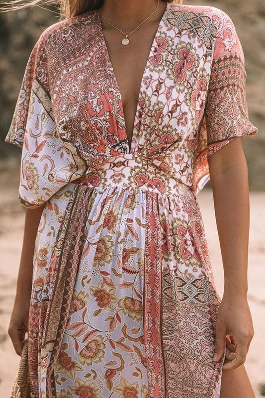 Multi - A woman on a sandy beach wearing a long, flowing pink and white bohemian print maxi dress with a deep V-neck, wide short sleeves, and a high leg slit, holding a woven straw bag.