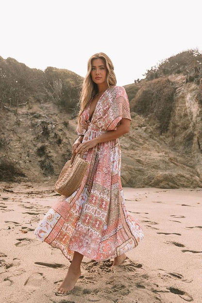 Multi - A woman on a sandy beach wearing a long, flowing pink and white bohemian print maxi dress with a deep V-neck, wide short sleeves, and a high leg slit, holding a woven straw bag.