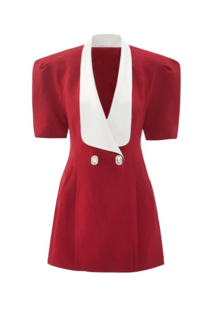 A woman with long blonde hair leans against a metal pole, wearing a structured red mini blazer dress with short puff sleeves, a wide white contrast lapel, and a flared A-line skirt.