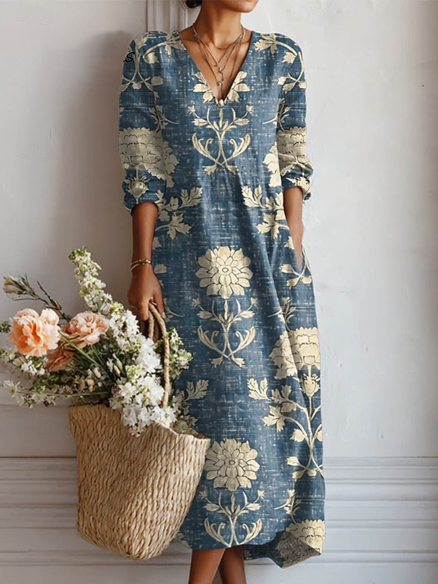 5XL - A woman wearing a blue slub-textured maxi dress with a cream botanical floral print, featuring a V-neckline, three-quarter sleeves, and side pockets, holding a straw bag with flowers.