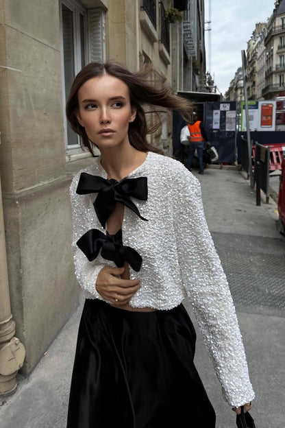 White - A model wearing a black cropped jacket covered in sequins, featuring two large black bows at the front closure.