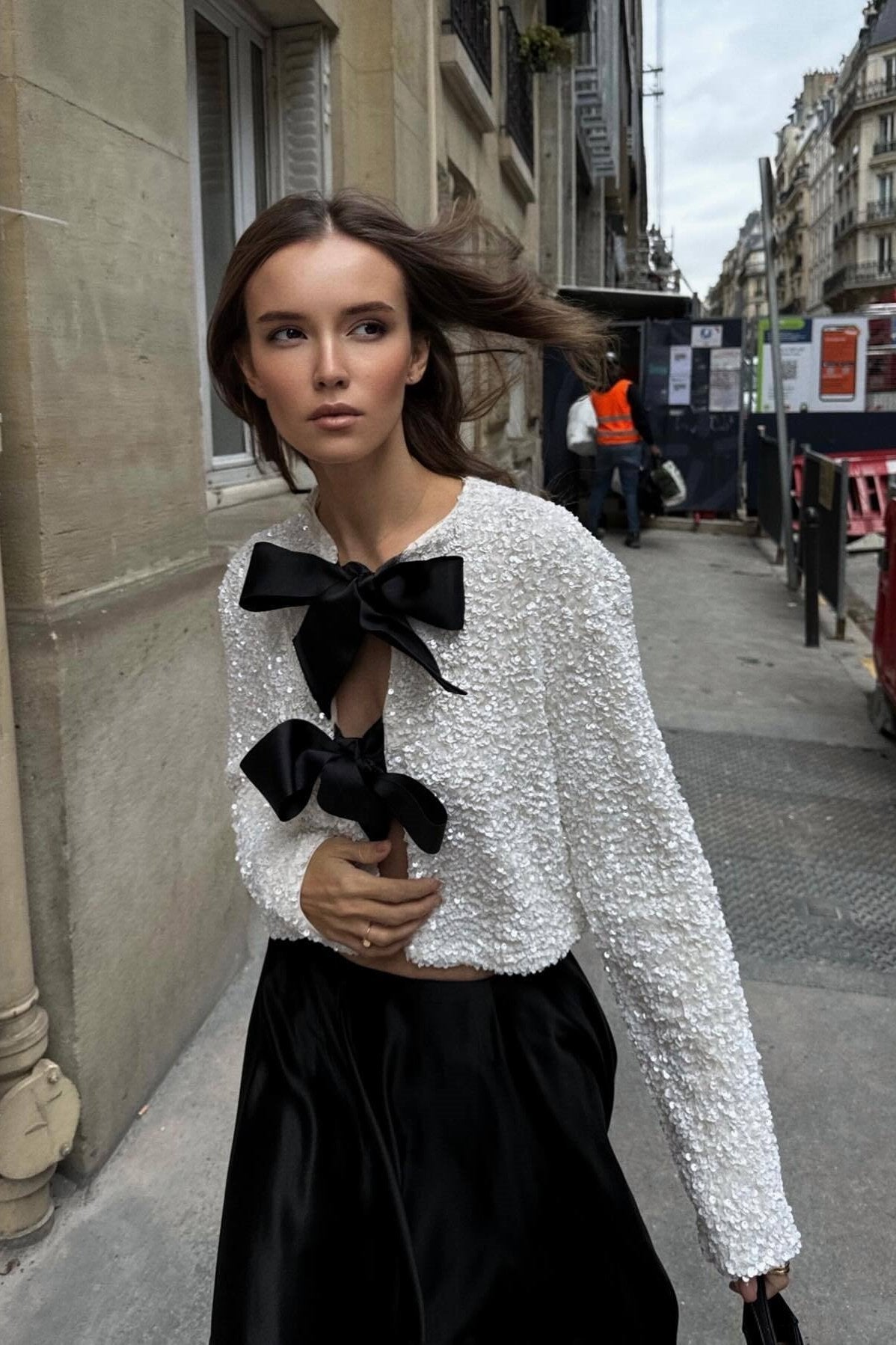 White - A woman wearing a black, long-sleeve cropped jacket covered in sequins, featuring two large black grosgrain bows tied at the front.