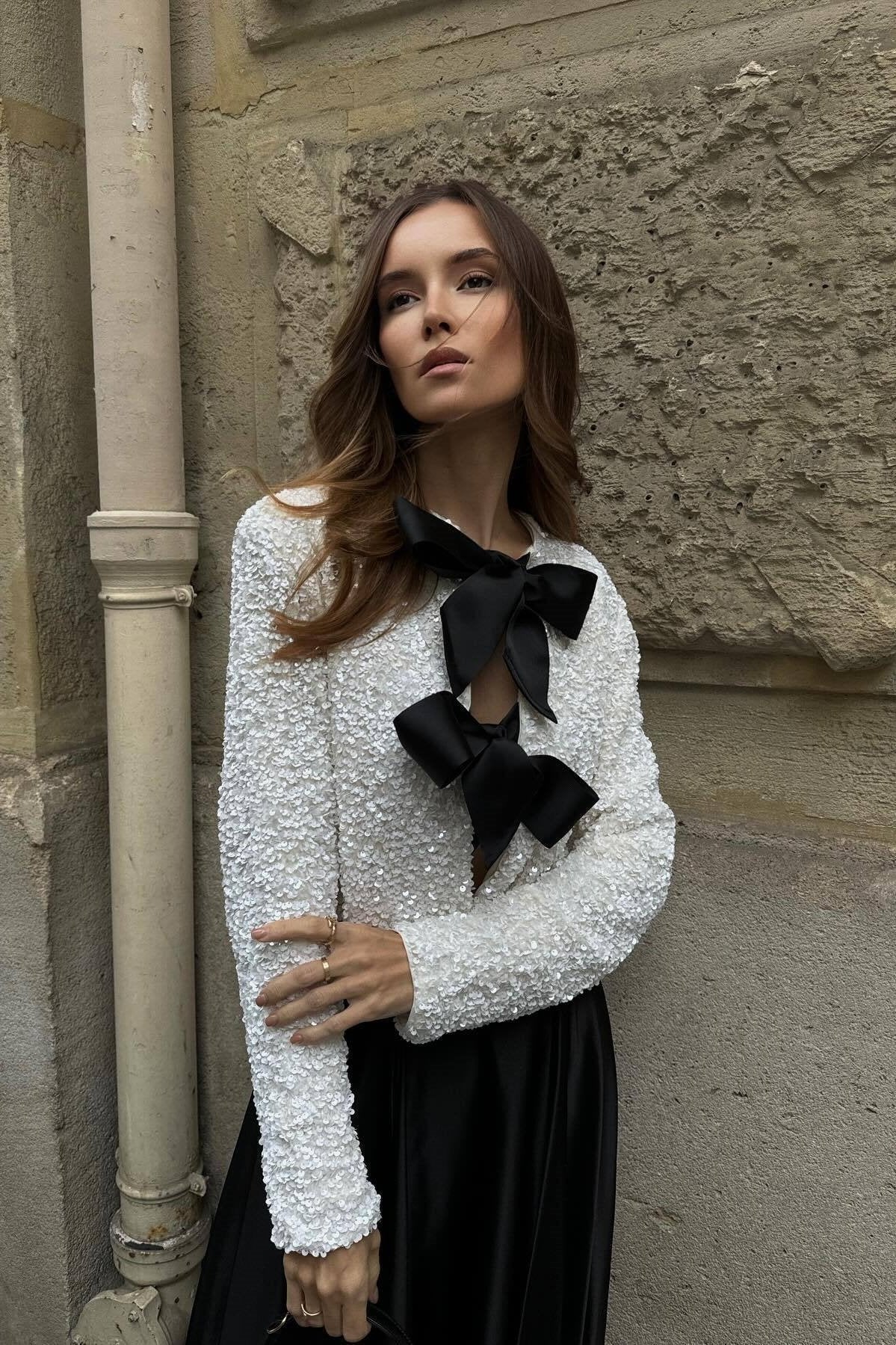 White - A model wearing a black cropped jacket covered in sequins, featuring two large black bows at the front closure.