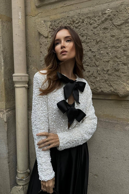 White - A model wearing a black cropped jacket covered in sequins, featuring two large black bows at the front closure.
