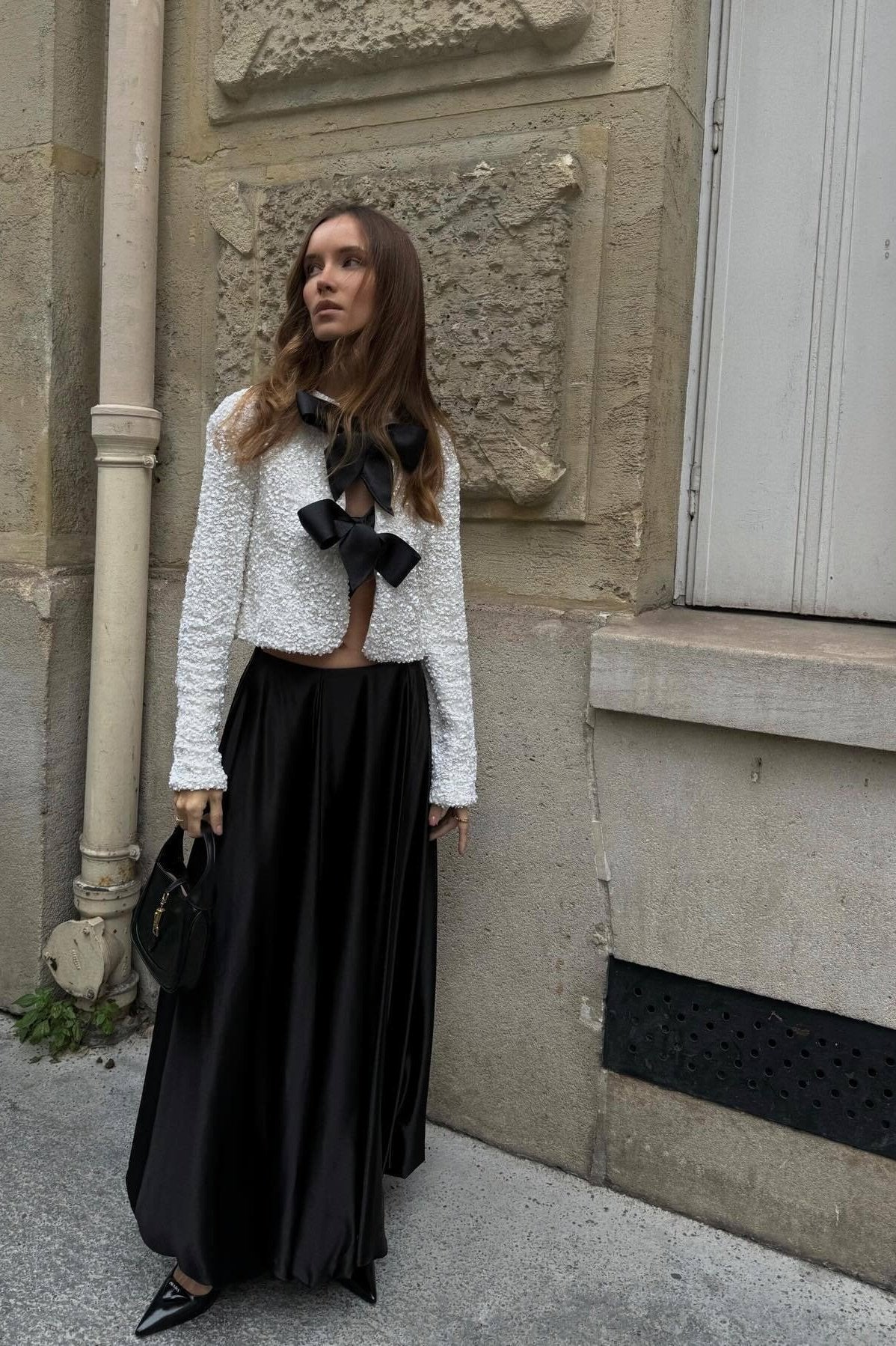 White - A woman wearing a black, long-sleeve cropped jacket covered in sequins, featuring two large black grosgrain bows tied at the front.