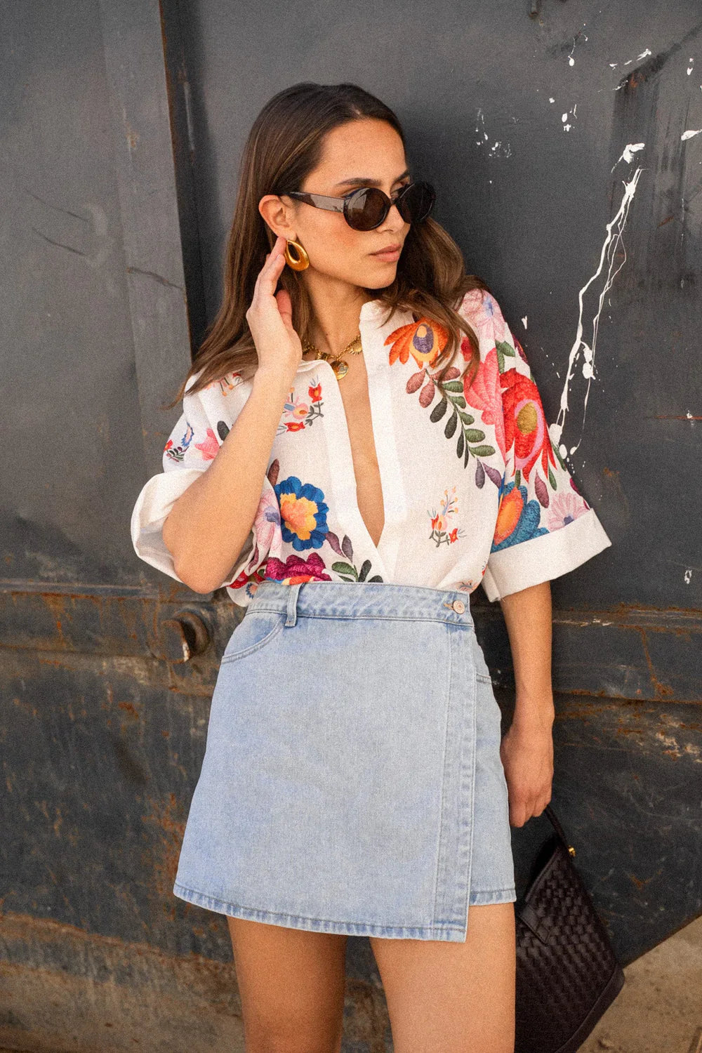 A woman wearing a white, short-sleeved button-down blouse adorned with vibrant, large-scale floral embroidery in shades of pink, blue, orange, and green.