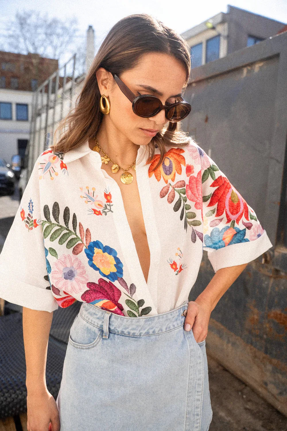 Print - A woman wearing a white, short-sleeved button-down blouse adorned with vibrant, large-scale floral embroidery in shades of pink, blue, orange, and green.