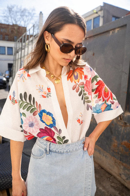 Print - A woman wearing a white, short-sleeved button-down blouse adorned with vibrant, large-scale floral embroidery in shades of pink, blue, orange, and green.