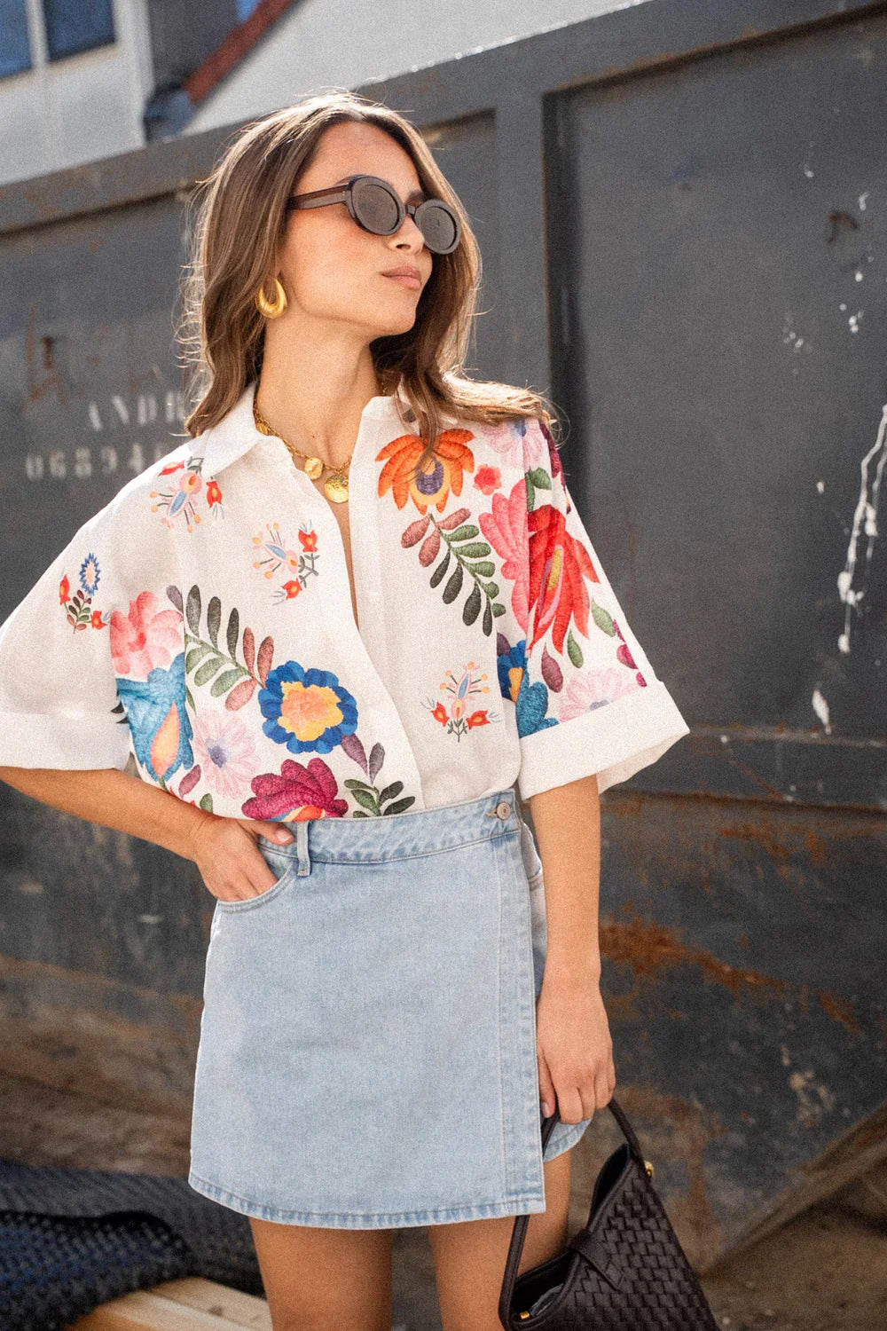 Print - A woman wearing a white, short-sleeved button-down blouse adorned with vibrant, large-scale floral embroidery in shades of pink, blue, orange, and green.