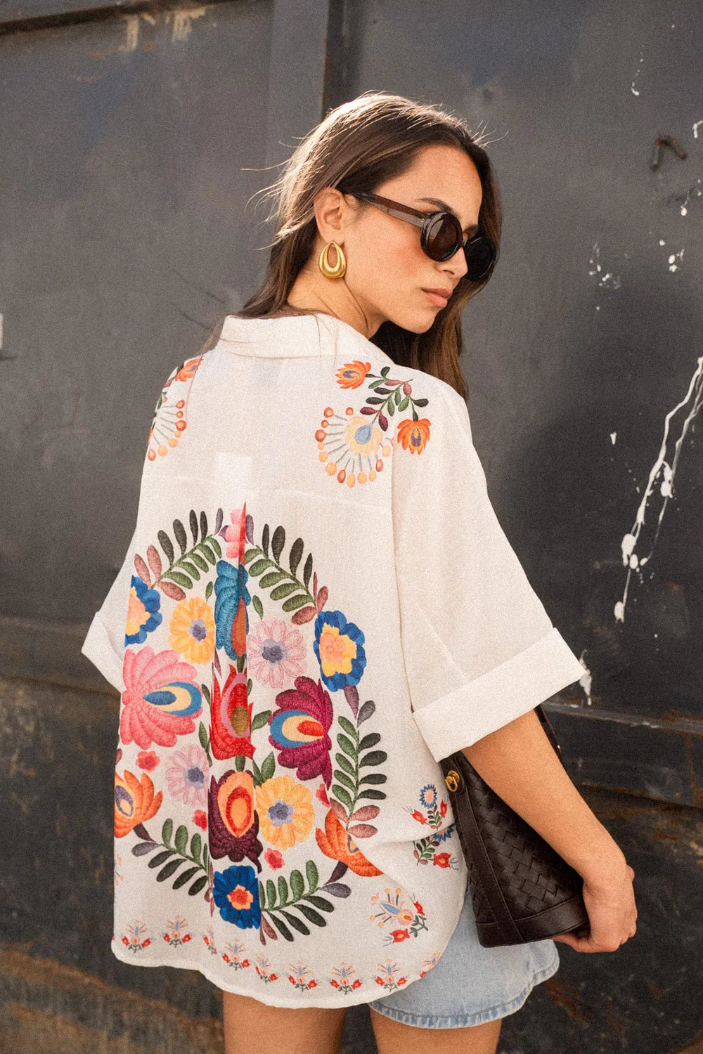 Print - A woman wearing a white, short-sleeved button-down blouse adorned with vibrant, large-scale floral embroidery in shades of pink, blue, orange, and green.
