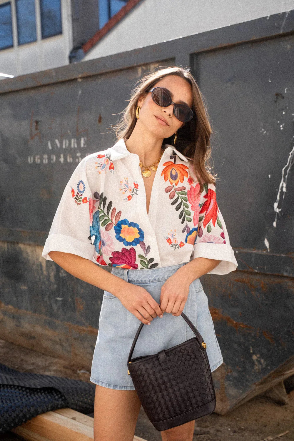 Print - A woman wearing a white, short-sleeved button-down blouse adorned with vibrant, large-scale floral embroidery in shades of pink, blue, orange, and green.
