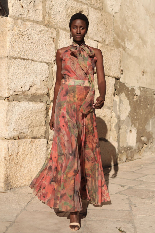 Flower - A model walking outdoors wearing a flowing, floral chiffon maxi dress featuring a halter neckline with an oversized bow and a banded waist.