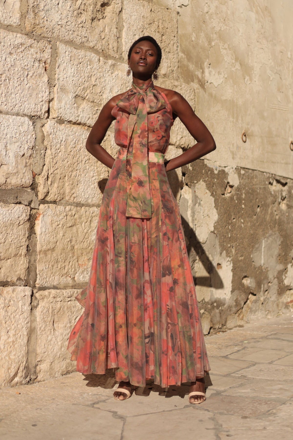 Flower - A model walking outdoors wearing a flowing, floral chiffon maxi dress featuring a halter neckline with an oversized bow and a banded waist.