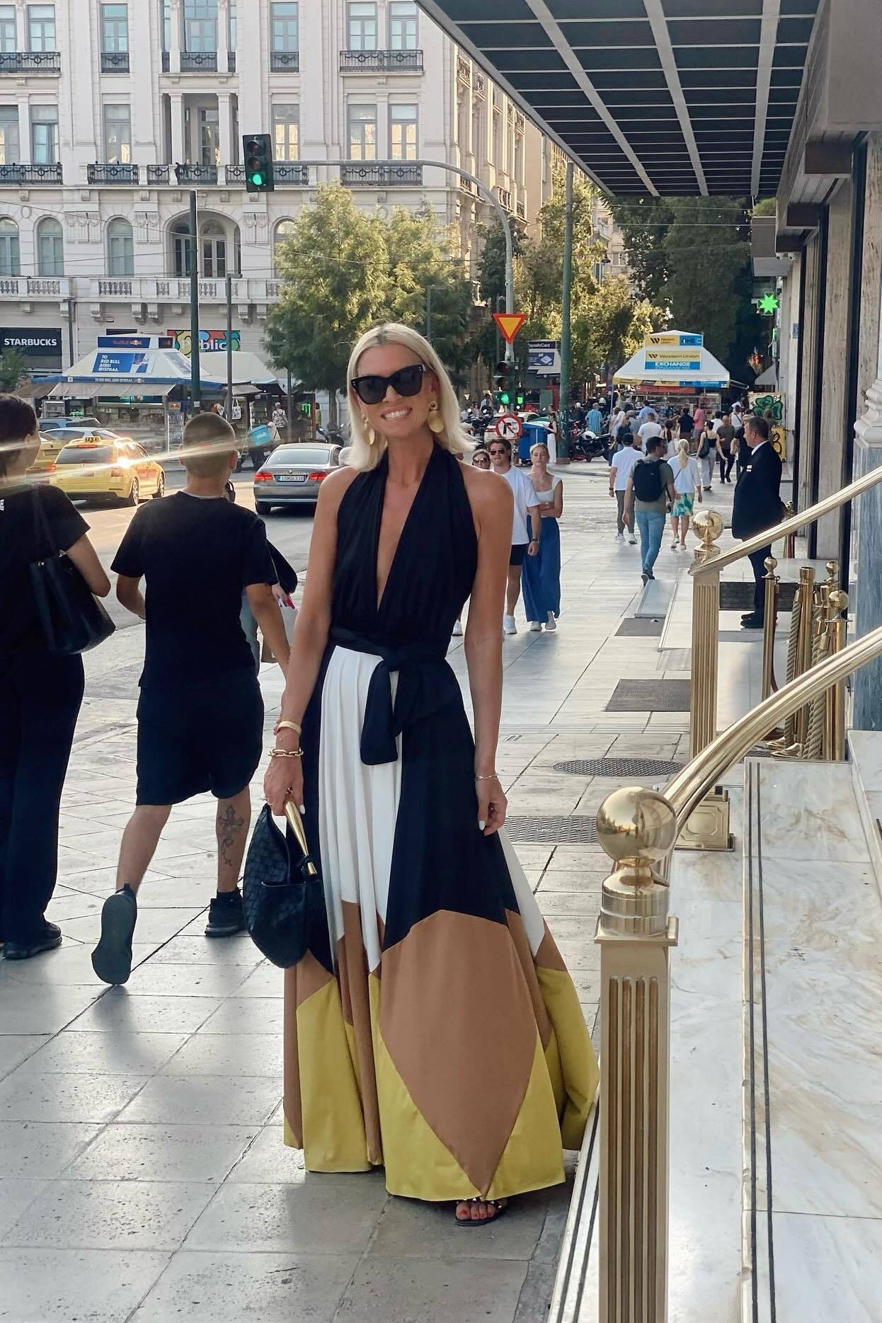 Multi - Woman wearing a plunging halter neck maxi dress with a black bodice, front tie, and a voluminous colorblock skirt in white, brown, and yellow, standing outdoors near palm trees.