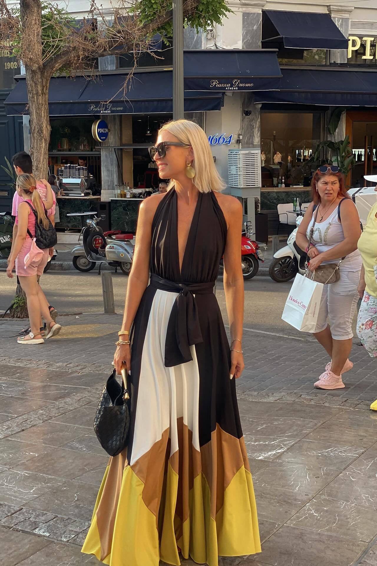 Multi - Woman wearing a plunging halter neck maxi dress with a black bodice, front tie, and a voluminous colorblock skirt in white, brown, and yellow, standing outdoors near palm trees.