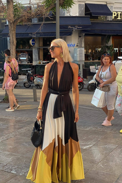 Multi - Woman wearing a plunging halter neck maxi dress with a black bodice, front tie, and a voluminous colorblock skirt in white, brown, and yellow, standing outdoors near palm trees.
