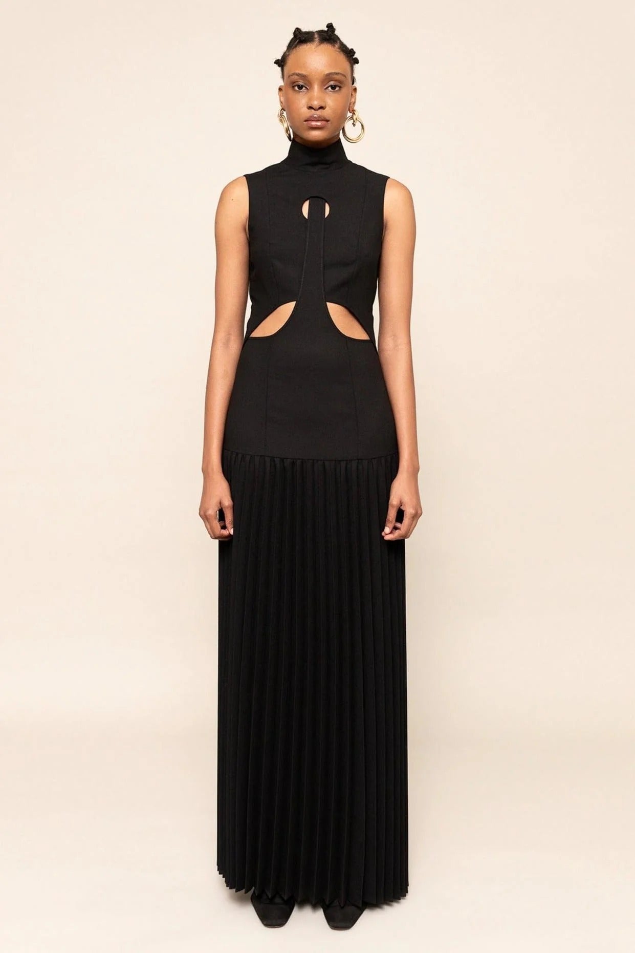 Black - A model wearing a sleeveless maxi dress featuring a high mock neck, geometric cutouts on the bodice, a drop waist, and a finely pleated floor-length skirt.