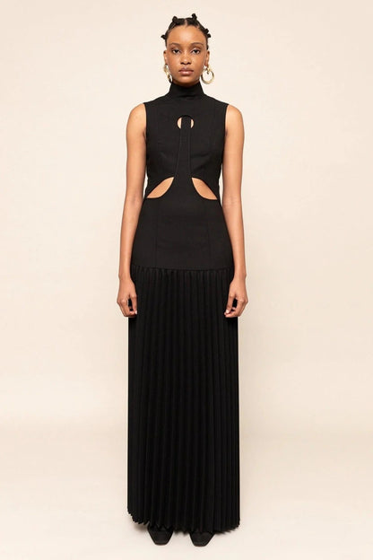 Black - A model wearing a sleeveless maxi dress featuring a high mock neck, geometric cutouts on the bodice, a drop waist, and a finely pleated floor-length skirt.