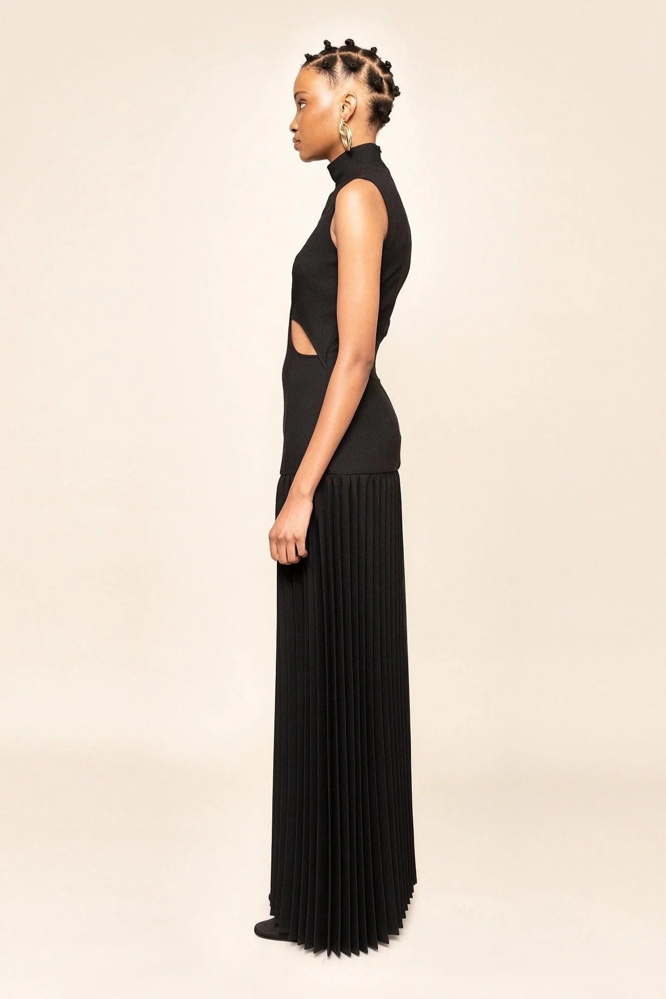 Black - A model wearing a sleeveless maxi dress featuring a high mock neck, geometric cutouts on the bodice, a drop waist, and a finely pleated floor-length skirt.