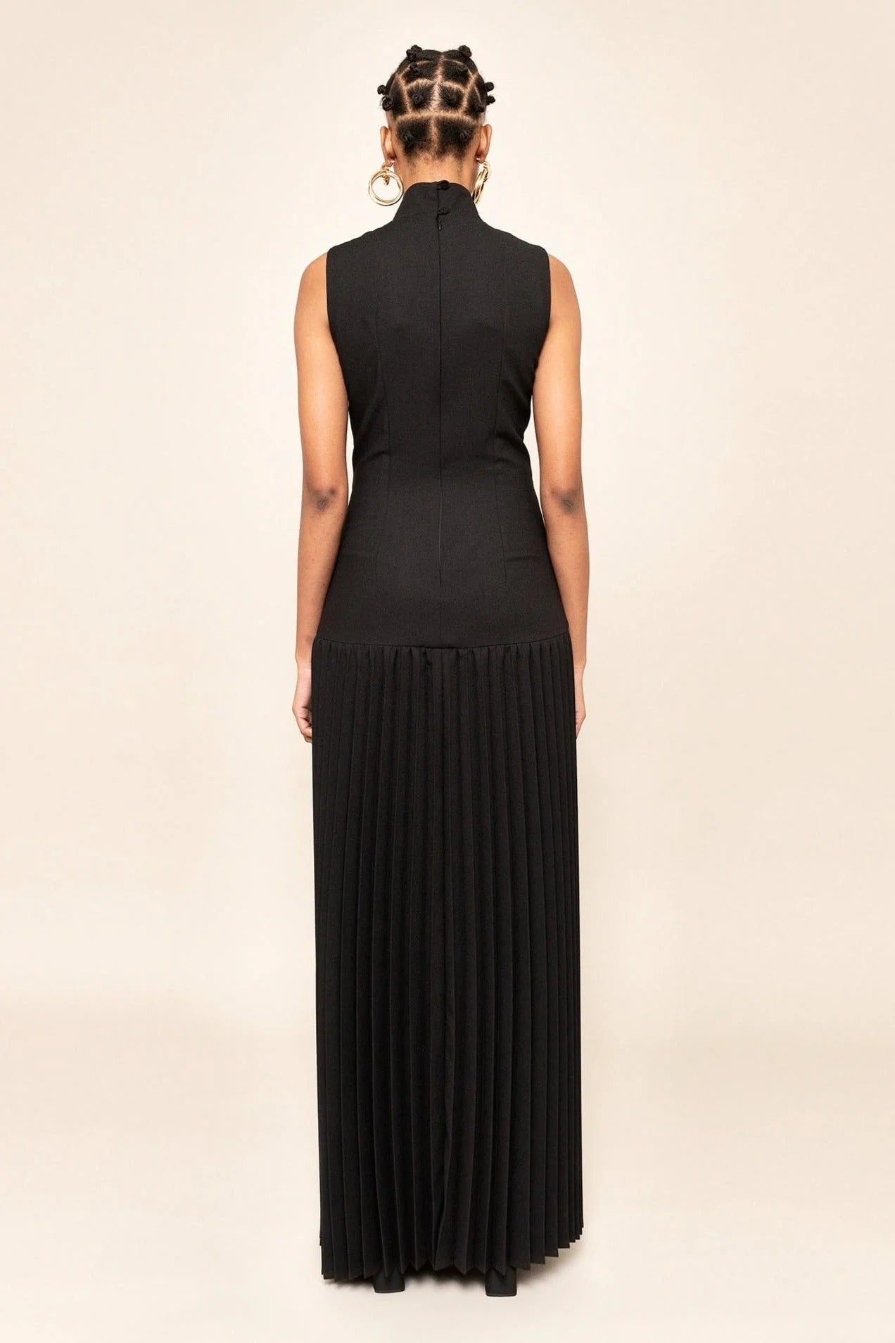 Black - A model wearing a sleeveless maxi dress featuring a high mock neck, geometric cutouts on the bodice, a drop waist, and a finely pleated floor-length skirt.