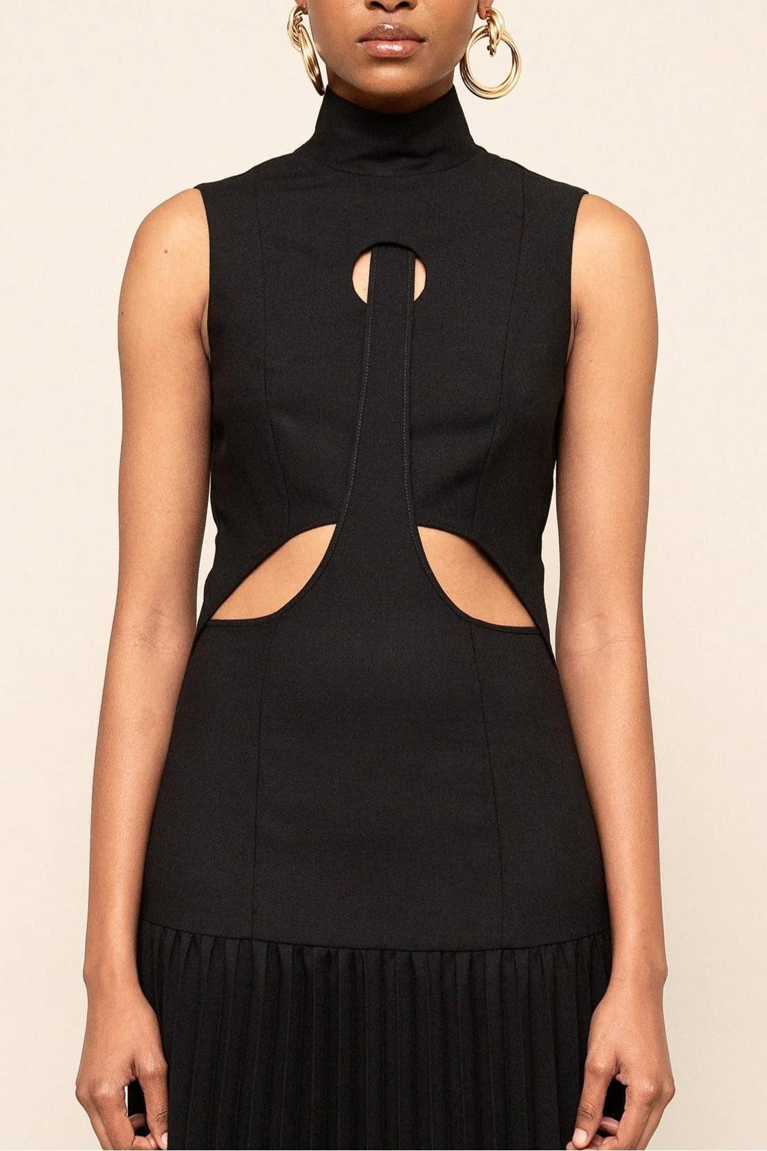 Black - A model wearing a sleeveless maxi dress featuring a high mock neck, geometric cutouts on the bodice, a drop waist, and a finely pleated floor-length skirt.
