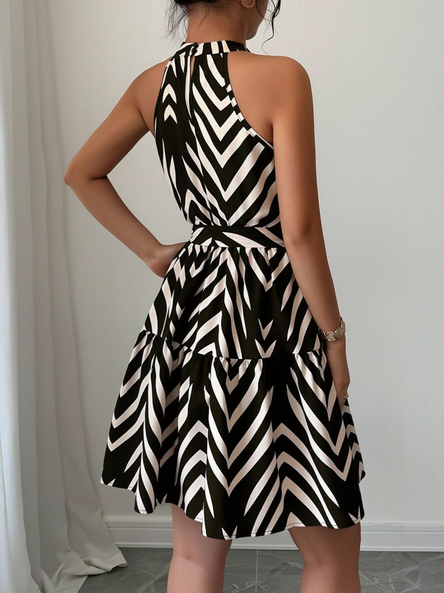 Black - Woman wearing a sleeveless halter neck mini dress with a geometric pattern, featuring a tie waist and tiered skirt