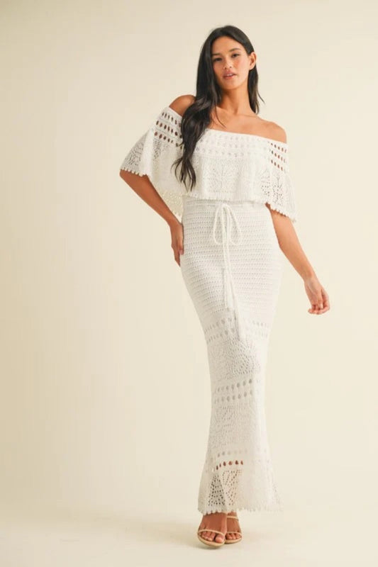 White - A woman wearing a white off-the-shoulder crochet maxi dress with a flounce bodice, a drawstring waist, and intricate open-knit patterns throughout.