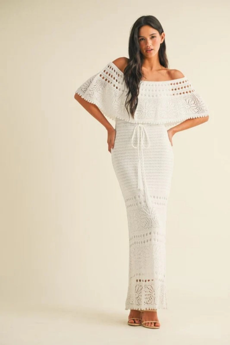 White - A woman wearing a white off-the-shoulder crochet maxi dress with a flounce bodice, a drawstring waist, and intricate open-knit patterns throughout.