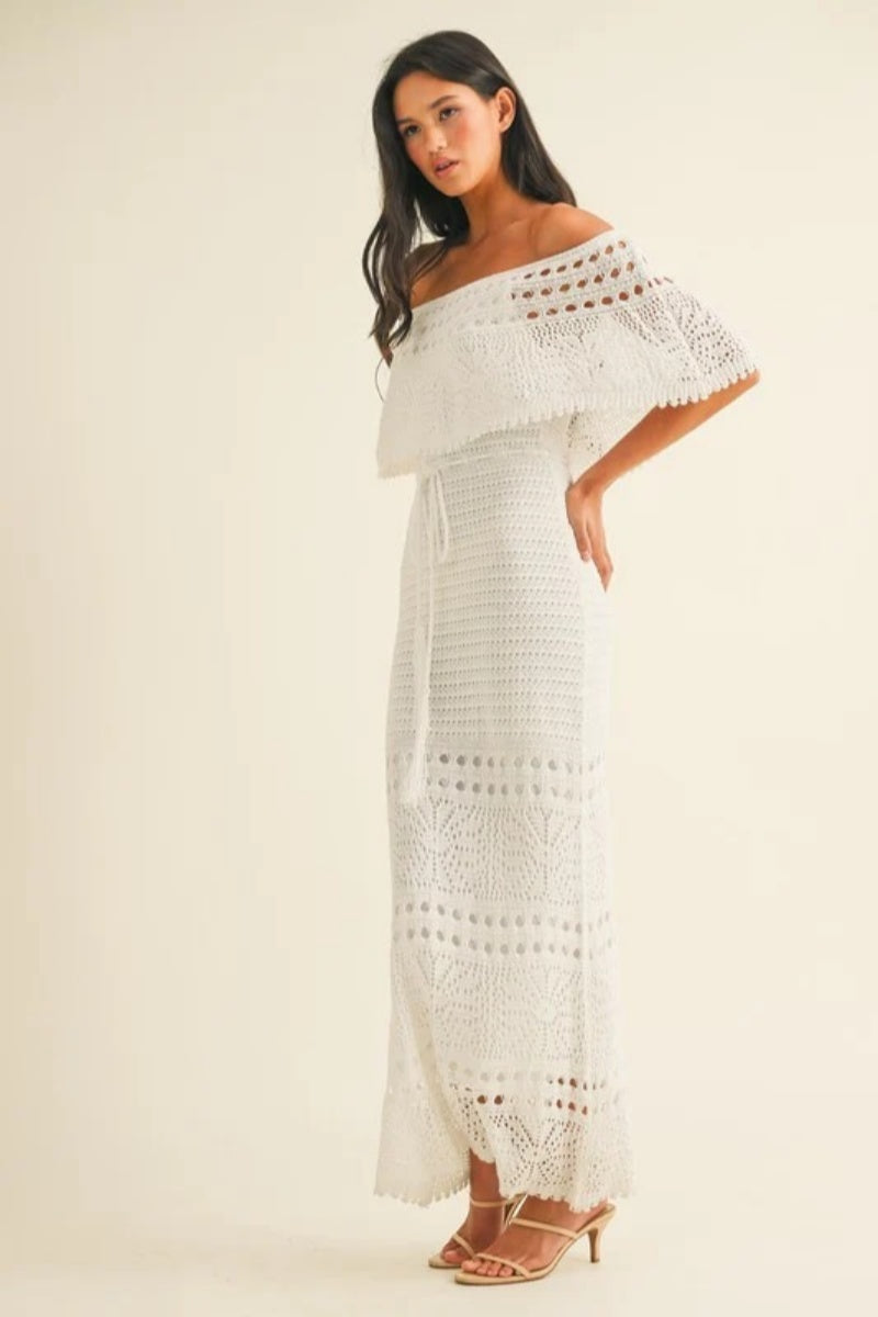 White - A woman wearing a white off-the-shoulder crochet maxi dress with a flounce bodice, a drawstring waist, and intricate open-knit patterns throughout.
